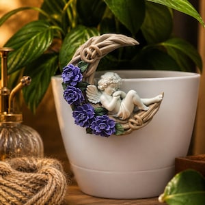 Könnte beinhalten: Ein weißer Keramik-Pflanztopf mit einer dekorativen Halbmond- und Engelsfigur. Der Mond ist mit lila Blumen geschmückt und der Engel liegt. Eine goldfarbene Pflanzenspritze und eine Schnurkugel befinden sich im Hintergrund.