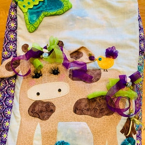 May include: A brown and white cow with purple and green ribbons, a yellow bird with a purple bow, and blue flowers on a green and white background. The cow has black eyes and a stitched nose. The background is a light blue and white fabric with a purple border.