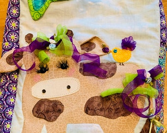 May include: A brown and white cow with purple and green ribbons, a yellow bird with a purple bow, and blue flowers on a green and white background. The cow has black eyes and a stitched nose. The background is a light blue and white fabric with a purple border.