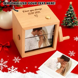 May include: A wooden music box with a photo of a couple kissing. The box has a crank and a small gold button. The top of the box has the text "I love you always 22.07.2025" and a heart symbol. The background is red with a small Christmas tree.