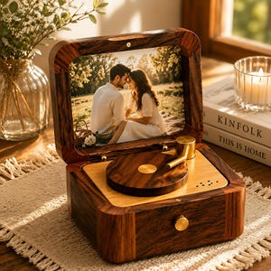 Puede incluir: Una caja de música de madera diseñada como un tocadiscos en miniatura con un marco de madera oscura. La tapa presenta una fotografía de una pareja sentada al aire libre. La base incluye un plato giratorio de madera, un brazo dorado y un pomo dorado.