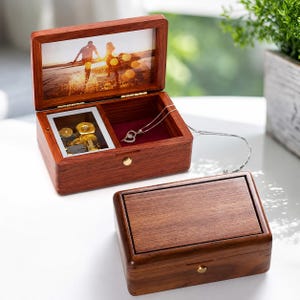 Könnte beinhalten: Eine rechteckige Spieluhr aus Holz mit dunkelrotbrauner Oberfläche. Der geöffnete Deckel zeigt ein Foto von zwei Personen am Strand. Im Inneren ist ein goldenes mechanisches Uhrwerk neben einem roten Samtfach mit einer silbernen Halskette zu sehen.
