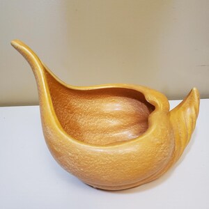 May include: A ceramic planter in a seashell design. The planter is a warm, speckled orange color with a textured surface. It features a curved, elongated neck and a flared, fan-shaped end. The interior is smooth and open.