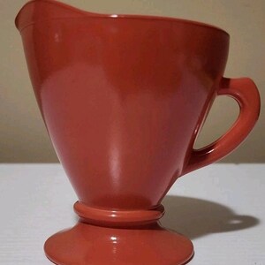 Op de afbeelding: Een rode creamer met een handvat en een uitlopende, conische vorm. De creamer heeft een ronde basis en een klein, gebogen handvat. De kleur is een effen, levendig rood en de creamer lijkt gemaakt van keramiek of een vergelijkbaar materiaal.