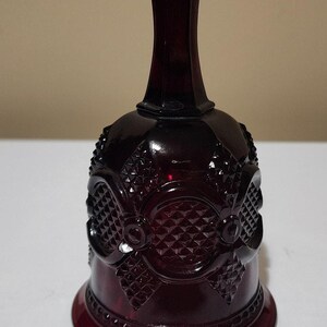May include: A ruby red glass bell with a detailed, textured pattern. The bell has a flared base, a rounded body with diamond and floral designs, and a long, slender neck leading to a rounded top. The bell is likely vintage.