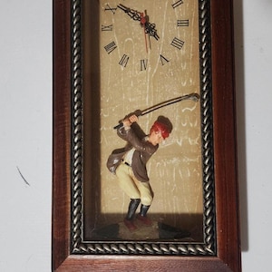 May include: A rectangular wall clock with a dark wood frame and a rope-textured inner border. The clock face features Roman numerals and a three-dimensional figure of a golfer in a brown jacket and tan pants swinging a golf club at the bottom.
