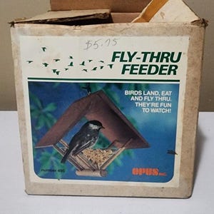 Puede incluir: Una caja de cartón para un comedero Fly-Thru Feeder de Opus Inc. La caja presenta un gráfico de un pájaro en un comedero de madera con el texto: Fly-Thru Feeder, Birds land, eat and fly thru. They're fun to watch! El número 495 está impreso.