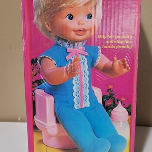 以下が含まれることがあります： ヴィンテージMattel「Bye Bye Diapers」人形の箱。箱はピンク色で、「Bye Bye Diapers」の文字が黄色で書かれています。人形は青い服を着て、ピンク色の便器に座っています。「Baby's a big girl now!」というテキストも表示されています。