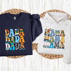 May include: A navy blue crew neck sweatshirt and a white hooded sweatshirt. Both sweatshirts feature colourful text designs. The navy sweatshirt says "DADA" in a stacked design. The white sweatshirt says "MAMA" in a stacked design.
