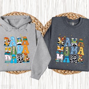 May include: Two grey sweatshirts, one a hoodie, featuring the words "DADA DADA DADA" and "MAMA MAMA MAMA" in colourful, cartoon lettering. The letters are filled with patterns and character designs, set against a light wooden background.