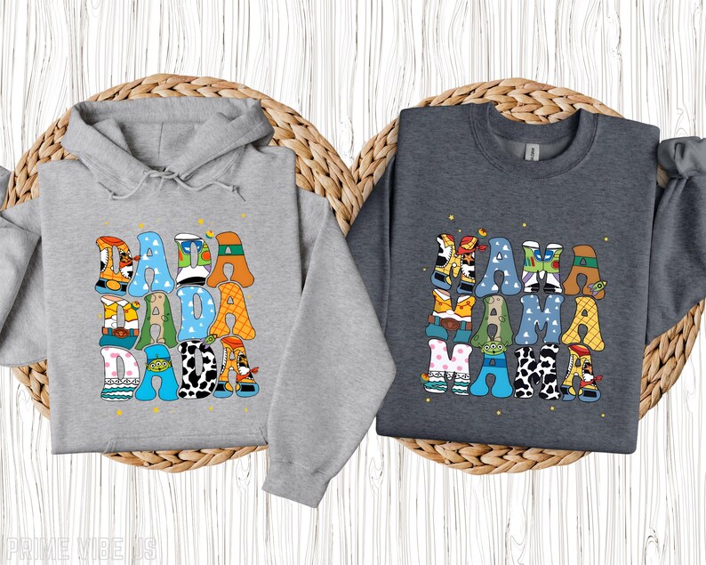 May include: Two grey sweatshirts, one a hoodie, featuring the words "DADA DADA DADA" and "MAMA MAMA MAMA" in colourful, cartoon lettering. The letters are filled with patterns and character designs, set against a light wooden background.