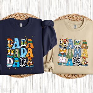May include: Two sweatshirts, one navy blue and one beige, each with a colourful graphic. The navy sweatshirt says "DADA" in large, playful letters, each decorated with a different pattern. The beige sweatshirt says "MAMA" in a similar style.