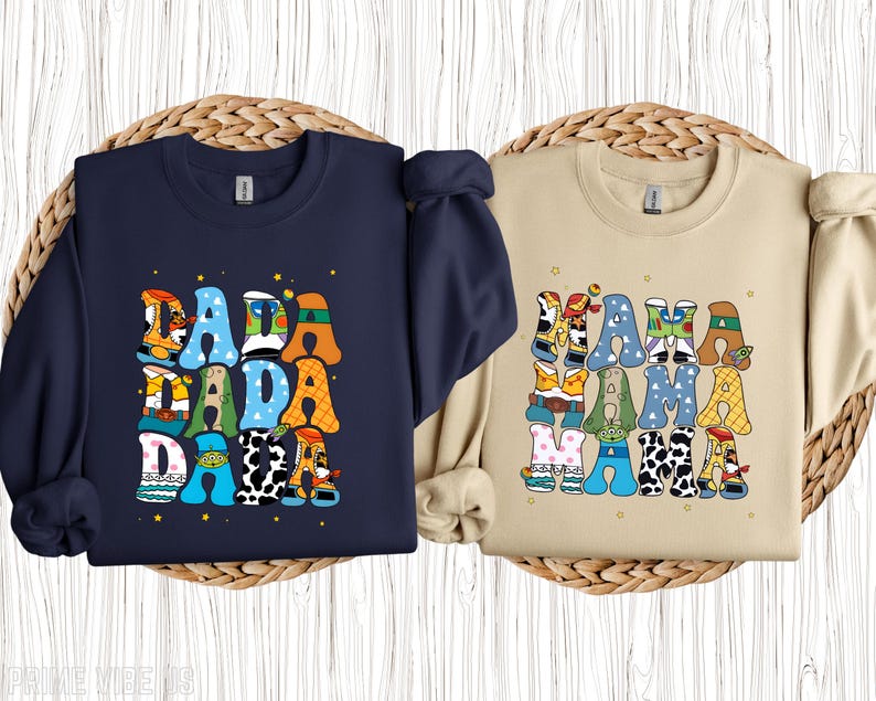 May include: Two sweatshirts, one navy blue and one beige, each with a colourful graphic. The navy sweatshirt says "DADA" in large, playful letters, each decorated with a different pattern. The beige sweatshirt says "MAMA" in a similar style.