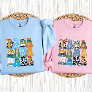 May include: Two crewneck sweatshirts, one light blue and one pink. The light blue sweatshirt has the word "DADA" in colourful, patterned letters. The pink sweatshirt has the word "MAMA" in colourful, patterned letters.