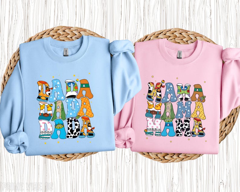 May include: Two crewneck sweatshirts, one light blue and one pink. The light blue sweatshirt has the word "DADA" in colourful, patterned letters. The pink sweatshirt has the word "MAMA" in colourful, patterned letters.