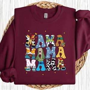 May include: A maroon sweatshirt featuring the word "MAMA" in a colourful, patterned design. The letters are themed around Toy Story, incorporating characters and elements from the film. Small yellow stars are scattered around the text.