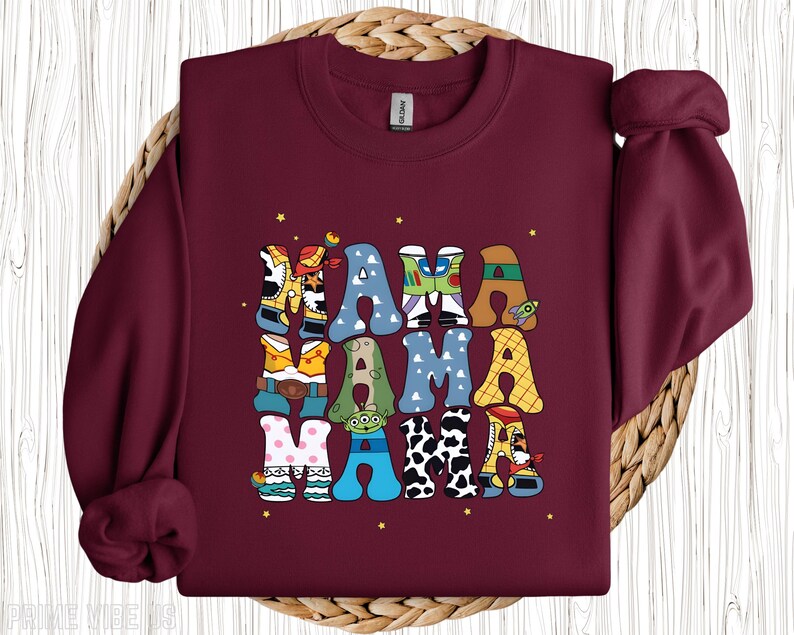 May include: A maroon sweatshirt featuring the word "MAMA" in a colourful, patterned design. The letters are themed around Toy Story, incorporating characters and elements from the film. Small yellow stars are scattered around the text.