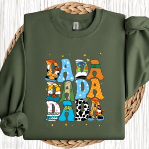 May include: An olive green sweatshirt featuring the word "DADA" in a playful, cartoon font. The letters are decorated with various patterns and character-themed designs, with small yellow stars dotted around the text.