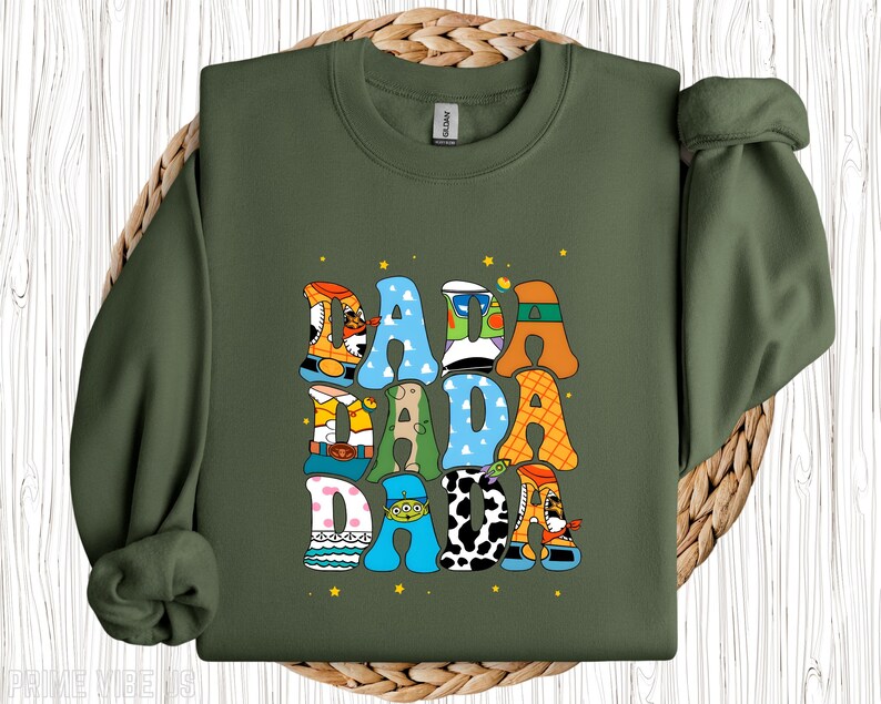 May include: An olive green sweatshirt featuring the word "DADA" in a playful, cartoon font. The letters are decorated with various patterns and character-themed designs, with small yellow stars dotted around the text.