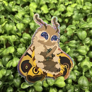 May include: A glittery sticker of a cartoon moth with yellow and black wings, holding a green leaf in its furry brown hands. The moth has large blue eyes and a pink blush on its cheeks.