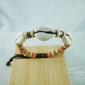 Puede incluir: Un brazalete de conchas blancas con cuentas de madera marrón y naranja. El brazalete está atado con un cordón negro.