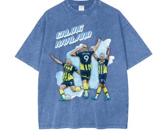 May include: A blue t-shirt with a faded look featuring a graphic of three soccer players in yellow and black uniforms. The text "ERLING HAALAND" is printed in white on the shirt.