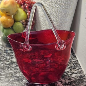 Könnte beinhalten: Ein roter Glaskorb mit einem Griff aus klarem Glas. Der Korb hat ein strukturiertes Aussehen und eine ausgestellte Form. Der Griff ist mit dekorativen Glasakzenten befestigt. Der Korb wird wahrscheinlich zu dekorativen Zwecken verwendet.