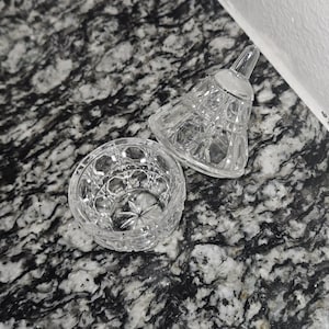 Puede incluir: Una campana de cristal tallado transparente con un cuenco abierto a juego. La campana está boca abajo, revelando sus intrincados patrones de diamantes y estrellas. El cuenco tiene un diseño de estrella en el centro. Los objetos están sobre una superficie moteada gris y blanca.