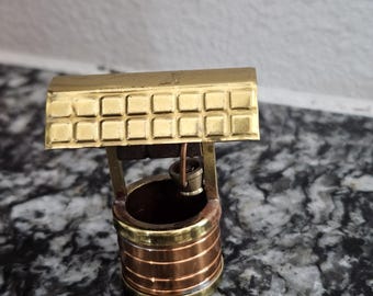 May include: A miniature wishing well crafted from polished brass and copper. The structure features a rectangular roof with a grid pattern, two vertical support posts, and a small bucket suspended by a thin wire inside the cylindrical base.