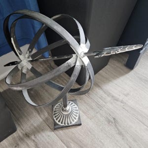 May include: A decorative metal sphere sculpture with a distressed gray finish. The sphere is supported by a pedestal base with a square bottom and a decorative, white, circular design. An arrow-shaped element extends from the sphere.