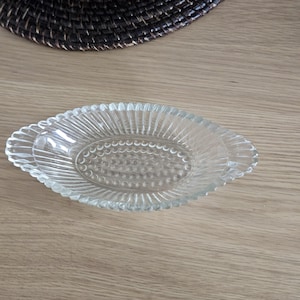 Puede incluir: Plato ovalado de vidrio transparente con un diseño texturizado y acanalado. El plato tiene un borde festoneado y un patrón central de pequeños puntos en relieve. El plato está sobre una superficie de madera clara.