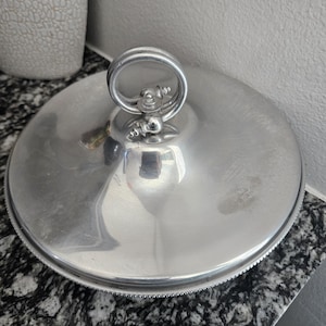 Puede incluir: Tapa redonda de metal plateado para servir, con asa de anillo. La tapa tiene un borde decorativo y una superficie reflectante. El asa está unida a la parte superior de la tapa con adornos decorativos.