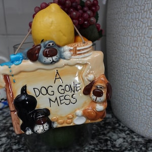 May include: A ceramic sign with a dog theme. The sign is brown and white with a blue and green border. The sign reads "A Dog Gone Mess". There are three cartoon dogs on the sign, one black, one brown, and one blue.