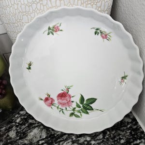 May include: White ceramic pie plate with a fluted edge. The plate is decorated with pink roses and green leaves. The roses are clustered in a corner and scattered around the plate. A decorative serving dish.