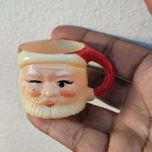 May include: A small, novelty Santa Claus mug with a red handle and hat trim. The mug features a winking Santa face with a white beard and rosy cheeks. The mug is made of a light-colored material.