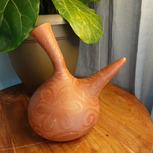 Puede incluir: Una jarra de terracota con un diseño único. La jarra tiene un cuerpo redondo con un pico largo y delgado y un asa. La jarra está decorada con un patrón repetitivo de flores estilizadas.