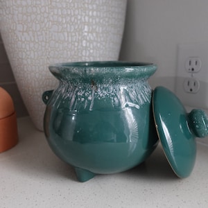 May include: A teal ceramic pot with a white glaze and a lid. The pot has three legs and a handle.
