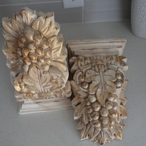 Puede incluir: Dos soportes de pared ornamentados de color blanco y dorado con un diseño de vid. Los soportes están hechos de madera y tienen un acabado desgastado.
