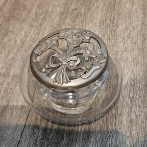 May include: A clear glass bowl featuring a circular silver-toned metal lid. The lid has a decorative cutout design depicting a bouquet of flowers tied with a ribbon. The bowl has a smooth, rounded base and a wide opening, providing a clear view.