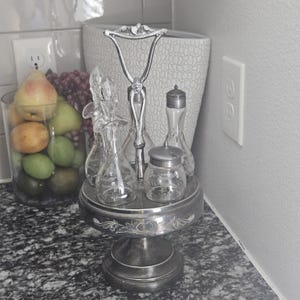 May include: A silver-toned condiment set with clear glass bottles and jars. The set includes a handle and sits on a decorative pedestal base. A glass jar of fruit is in the background.