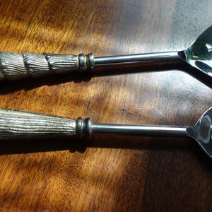 Op de afbeelding: Een opscheplepel en vork set. De handvatten hebben een zilverkleurig, getextureerd ontwerp en de lepels en vorken zijn glanzend. De set staat op een houten oppervlak. De handvatten hebben een houtachtige textuur.