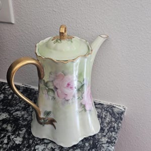 Puede incluir: Una tetera de porcelana vintage con un acabado verde claro y dorado. La tetera presenta una tapa y un asa con detalles dorados. Rosas rosas están pintadas en el cuerpo de la tetera, agregando un toque floral al diseño.