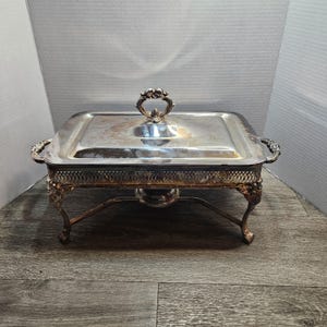 May include: A rectangular silver-plated chafing dish featuring a removable lid with a decorative handle, ornate side handles, and four intricate legs. The base of the dish has a lattice-style gallery border and a central support stand.