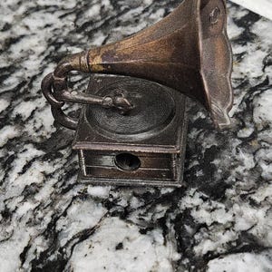 May include: A miniature antique-style gramophone pencil sharpener. The pencil sharpener is made of a dark, metallic material with a detailed design, including a horn, turntable, and a small hole for sharpening pencils.