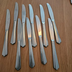 Puede incluir: Un conjunto de diez cuchillos de mesa de acero inoxidable con un diseño de mango acanalado. Los cuchillos están dispuestos sobre una superficie de madera, mostrando sus hojas pulidas y sus mangos decorativos. Los cuchillos son para cortar alimentos.