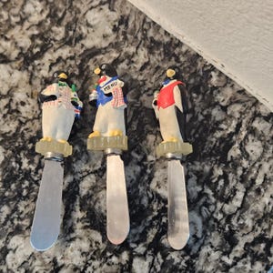 Può includere: Tre spalma formaggio decorativi con figurine di pinguini. Ogni pinguino indossa un abito diverso, con corpi bianchi e neri e accessori colorati. Gli spalma formaggio hanno lame argentate e sono su una base chiara.