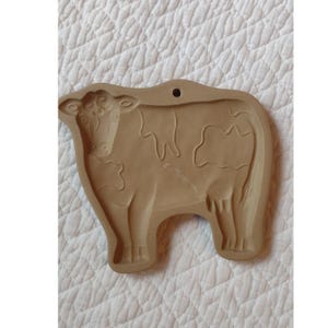 May include: Tan-colored cow-shaped cookie cutter with a hole at the top for hanging. The cow has detailed features, including spots and facial features. The cookie cutter is made of a durable material.