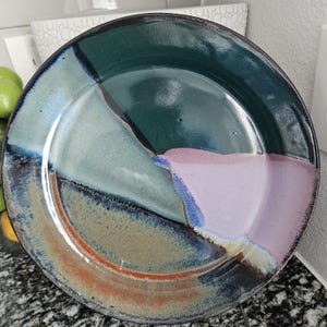 Puede incluir: Un plato de cerámica hecho a mano con un diseño abstracto único. El plato presenta una combinación de colores, incluyendo verde, azul, rosa y marrón. El borde del plato es oscuro y el interior está dividido en secciones de color.