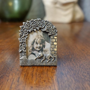 Könnte beinhalten: Ein silberfarbener Bilderrahmen mit floralem Design und einem Ziegelrand. Der Rahmen ist für ein 1 1/2 Zoll großes Foto ausgelegt. Der Rahmen hat unten eine kleine, silberfarbene Tierfigur.
