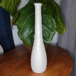 Könnte beinhalten: Weiße Keramikvase mit einem strukturierten Blasendesign. Die Vase ist hoch und schmal mit einem abgerundeten Boden.